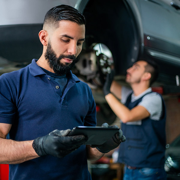 Supervisor at a car workshop checking tablet while mechanic works at background on a car